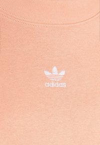 Närbild av ett persikofärgat tyg med en vit broderad Adidas trefoil-logotyp och text på bröstområdet.