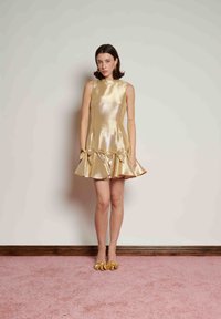 Χρυσό μεταλλικό φόρεμα σε γραμμή Α με ψηλό λαιμό, βολάν στο τελείωμα και φιόγκους στη μέση· συνοδεύεται από παπούτσια με χρυσές διακοσμήσεις.