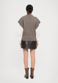 Frau steht mit dem Rücken zur Kamera und trägt einen braunen Strickwesten über einer weißen Langarmbluse, einen dunklen Leder-Minirock und braune Stiefeletten.