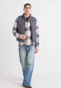 Chaleco acolchado gris sobre una camisa de cuadros con botones, combinado con jeans de un azul claro. Diseño simple, corte relajado y patrones mixtos.