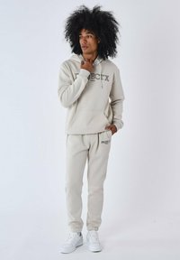 Sudadera beige con logo, bolsillo tipo pouch y cordón. Pantalones de chándal a juego con puños elásticos. Combinado con zapatillas blancas sobre un fondo liso.