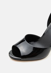 Μαύρο ψηλοτάκουνο παπούτσι από δερματίνη με ανοιχτή μύτη και λουράκι στον αστράγαλο, εμφανισμένο πάνω σε απλό φωτεινό φόντο.