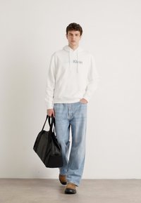 Biała bluza z kapturem i sznurkiem oraz logo, jasnoniebieskie dżinsy, czarna torba sportowa, brązowe buty, stojący na jednolitym tle.