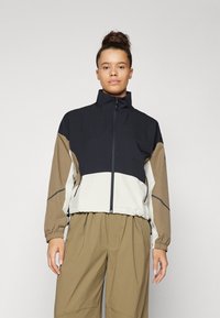Spalvotai blokuota zip-up striukė juodos, kreminės ir smėlio spalvos; turi elastingus rankogalius ir aukštą apykaklę; pagaminta iš lengvos, tekstūruotos medžiagos.