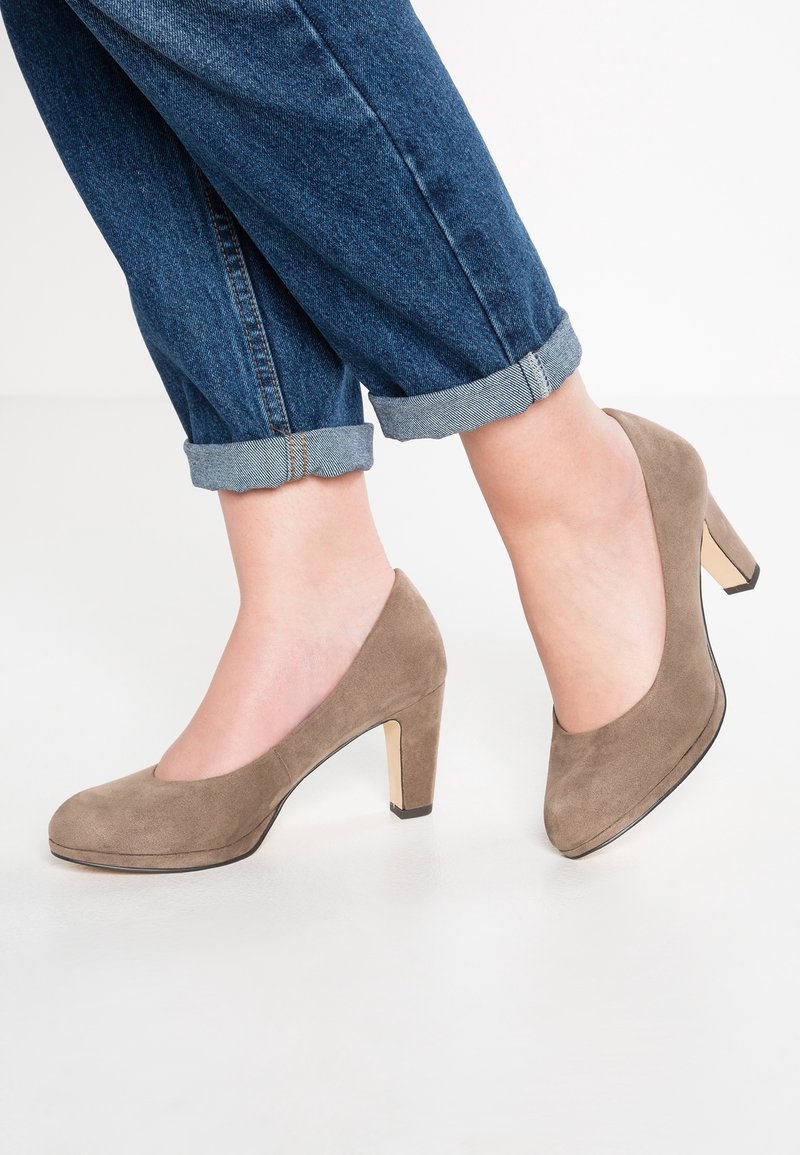 Femme portant un jean bleu retroussé et des escarpins en daim taupe à talon moyen, debout sur un sol blanc.