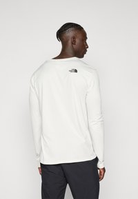 Man with braided hair wearing a white long-sleeve shirt with The North Face logo on upper back and black pants, facing sideways.