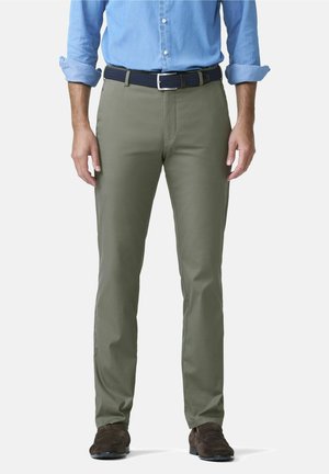 Mann trägt olivgrüne Hose, dunklen Gürtel, blaues Hemd mit aufgekrempelten Ärmeln und braune Schuhe, steht vor weißem Hintergrund.