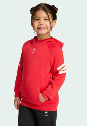 Ragazza sorridente, con le mani in tasca, che indossa una felpa rossa con cappuccio e pantaloni neri con i loghi Adidas e Disney su sfondo chiaro.