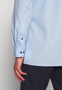 Lichtblauwe langemouwen shirt met knoopmanchetten, met een gladde textuur en twee zwarte knopen op elke manchet, getoond met een hand aan de zijkant.