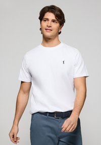 Witte katoenen t-shirt met korte mouwen en een ronde hals. Voorzien van een klein marineblauw logo op de borst. Gecombineerd met blauwe broek en een geweven riem.
