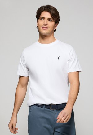 T-shirt bianco in cotone con maniche corte e collo rotondo. Presenta un piccolo logo blu navy sul petto. Abbinato a pantaloni blu e una cintura intrecciata.