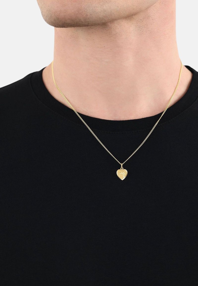 Gul hjärtformat hänge på en fin kedja, bärs över en svart t-shirt. Hänget har en intrikat design och polerad yta.