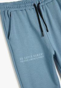 Lyseblå sweatpants lavet af blødt stof, med en elastisk talje og en sort snor, samt trykt tekst på låret.