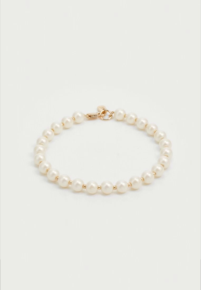 Un bracelet de perles couleur crème avec de petits intercalaires dorés et un fermoir doré, disposé en ovale sur un fond clair uni.