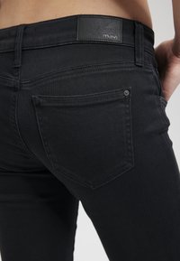 Musta farkkutakki, jossa on mavi-merkin nahkalaminaatti, näkyvät takataskut, sileä pinta ja slim-fit-muotoilu.