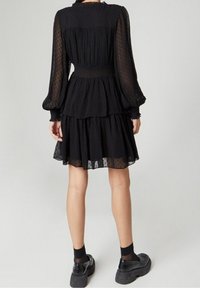 Svart klänning med genomskinlig prickig tyg, samlad midja och lager på lager kjol. Långa puffärm med elastiska manschetter. Buren med kraftiga svarta skor.