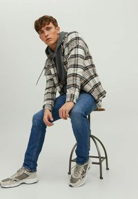 Camisa de cuadros en beige, negro y blanco sobre una sudadera gris, combinada con jeans de mezclilla azul y zapatillas grises, sentado en un taburete de metal.