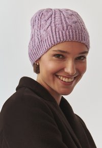 Lillasest lõngast kootud beanie, millel on soonikujuline tekstuur ja nöörkudumismuster. Mudel kannab tumedat jakki ja tal on lühikesed juuksed.