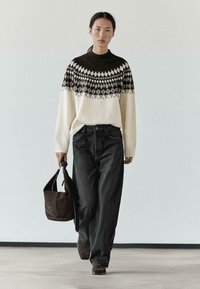 Woman walking in fair isle sweater, black wide-leg jeans, dark boots, and carrying a large dark suede handbag against a plain wall.