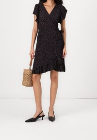 Abito nero avvolgente con motivo floreale, maniche corte a sbuffo, orlo arricciato, abbinato a una borsa intrecciata e tacchi a fascia neri.