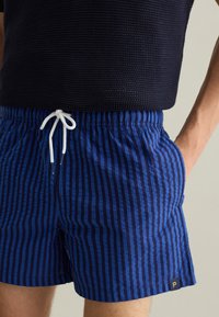 Costumi da bagno a righe blu con vita regolabile tramite cordoncino, tasche laterali e una piccola etichetta con logo. Il tessuto ha una consistenza morbida con linee verticali.