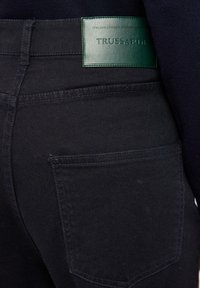 Černé džínové kalhoty s hladkým povrchem, s zelenou koženou etiketou označenou "TRUSSARDI" na zadním pasu a jednou zadní kapsou.