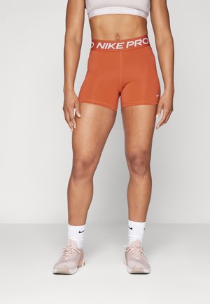 Fit Person in rostfarbenen Nike Pro Shorts, weißem Nike Crop-Top, weißen Nike Socken und pinken Sportschuhen, stehend vor einem schlichten Hintergrund.