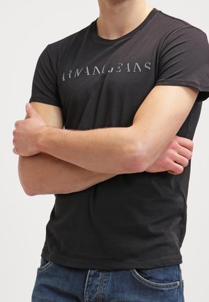 Homme les bras croisés portant un t-shirt noir Armani Jeans et un jean en denim bleu sur un fond uni.