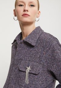 Lilla herringbone-mønstret jakke med klassisk krave og et stjerneformet broche med hængende rhinsten på lommen.