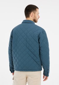 Gequilte blauwe jas met een ruitpatroon, een spitse kraag en verstelbare manchetten. Het materiaal lijkt zacht en lichtgewicht te zijn.