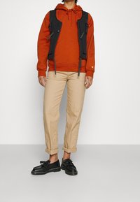 Chaleco oscuro sobre una sudadera naranja, pantalones beige y zapatos negros con borlas. Los detalles notables incluyen los bolsillos y los cordones del chaleco.