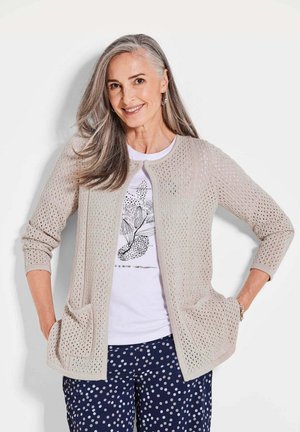 Cardigan beige en tricot avec un tissage ouvert texturé. Il possède de longues manches, deux poches avant et un col rond. Porté par-dessus une chemise blanche et un pantalon à pois foncés.