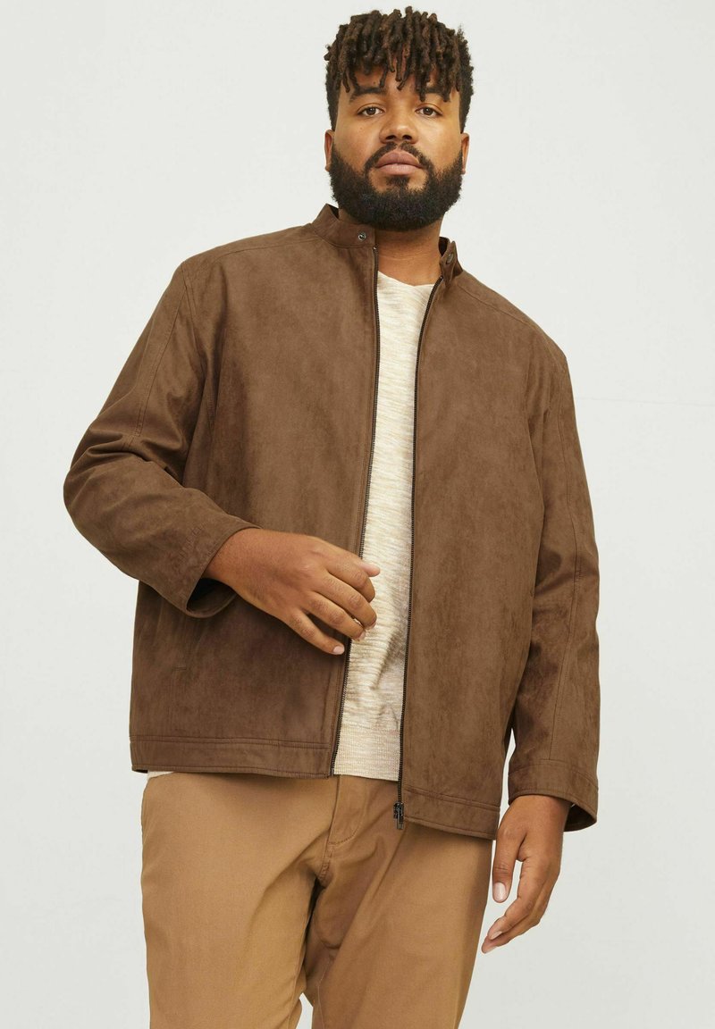 Chaqueta de ante marrón con un cuello alto y cierre de cremallera; presenta mangas largas y un ajuste relajado, combinada con una parte superior de color claro.