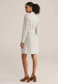 Vestito grigio chiaro a maniche lunghe con colletto e cintura in vita. Il tessuto ha una consistenza morbida; lunghezza al ginocchio con taglio dritto. Scarpe con tacco marroni.