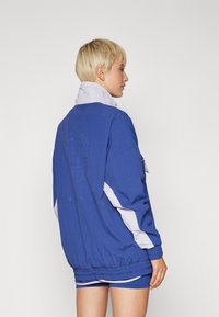 Person mit kurzem blonden Haar, der eine lockere blau-weiße Jacke und passende blaue Shorts trägt und seitlich vor einem einfarbigen Hintergrund steht.