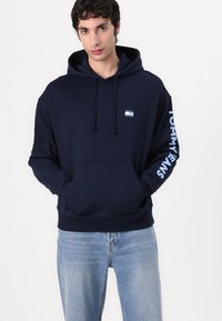 Sudadera azul marino con un bolsillo frontal, capucha con cordón y texto en blanco "TOMMY JEANS" en la manga izquierda. El material parece suave y cómodo.