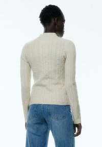 Cremfarbener Zopfmuster-Pullover mit Rundhalsausschnitt und langen Ärmeln, gefertigt aus strukturiertem Strickstoff, kombiniert mit hochtaillierten blauen Jeans.
