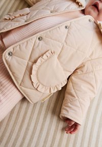 Veste crème matelassée avec poche à volants, fermetures éclairs argentées et texture douce, posée sur un fond en tissu rayé.