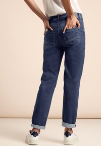 Dark blue denim jeans with a relaxed fit, featuring rolled cuffs, two back pockets, and subtle stitching details. Worn with white sneakers.