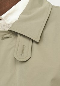 Světle béžové trench coat límec s jemnou texturou. Značné švy a kovové detaily na límci. Hladký povrch látky.