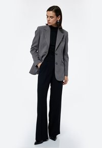 Kvinde iført grå blazer over sort ribbet turtleneck og vide sorte bukser, stående med den ene hånd i lommen foran en hvid baggrund.
