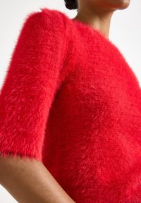 Maglione rosso morbido con maniche corte, superficie texturizzata e vestibilità rilassata, mostrato da un'angolazione bassa che mette in evidenza la morbidezza e il colore.