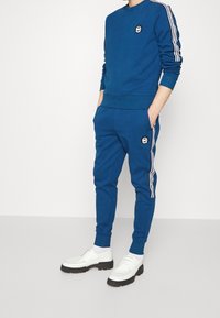 Felpa blu e pantaloni coordinati realizzati in morbido materiale, con strisce bianche sui lati e toppe logate sul petto e sulla coscia.
