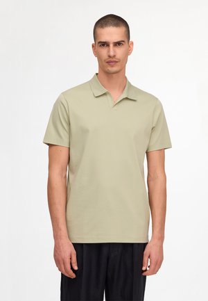 Mężczyzna z krótkimi włosami, ubrany w jasnozieloną koszulkę polo z krótkim rękawem i czarne spodnie, stojący na jednolitym białym tle.