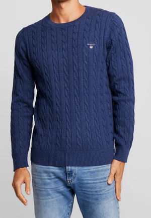 Homme portant un pull bleu GANT en maille torsadée avec poignets et ourlet côtelés, associé à un jean bleu clair.