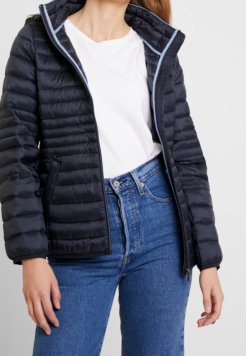 Doudoune navy avec une texture matelassée, accents bleu clair le long de la fermeture éclair et du col, associée à un jean en denim bleu taille haute.