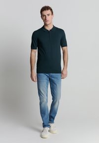 Donkergroen polo met korte mouwen en een ritskraag, gedragen met lichtblauwe jeans en witte sneakers, staande tegen een neutrale achtergrond.