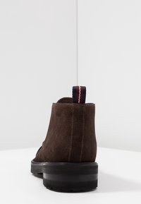 Bruine suede enkelboot met een hoge achterkant en een zwarte rubberen zool. Heeft een gestreepte trekband in marineblauw en bordeaux aan de hak.