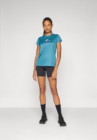 Teal athletic t-shirt with short sleeves and a mountain graphic, paired with black shorts. Model wears black socks and sneakers.