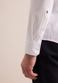 Zbliżenie na męski rękaw białej koszuli z czarnym haftem w kwiaty i zapięciem na guzik, na neutralnym tle.